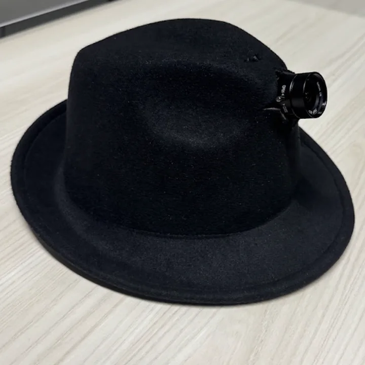 The hat by itself on a desk, from its right: a black felt hat, the lens standing out of the front of the crown, pointing right.
