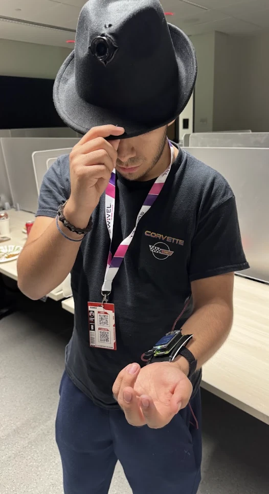 A crew member tips the hat. Its camera sits in the crown, and the wrist unit is strapped to the other forearm.