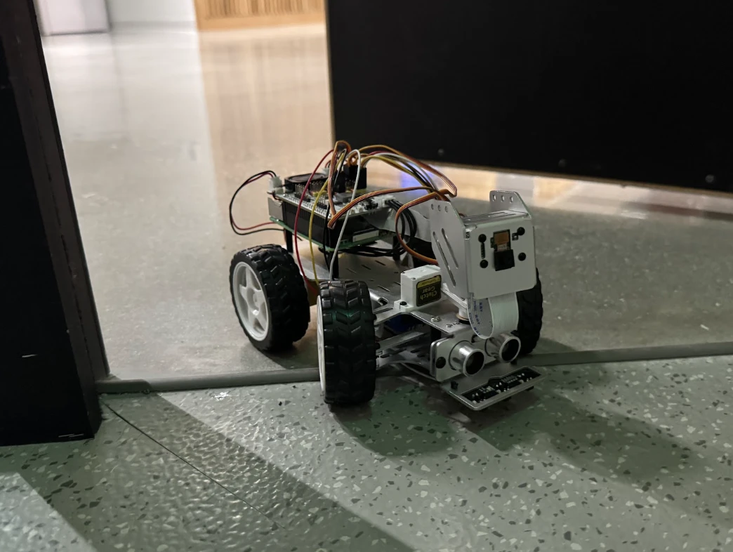 The rover on a speckled floor at an open doorway, seen from low down: camera and range sensors forward, a cable strip under its front wheels, a corridor beyond.