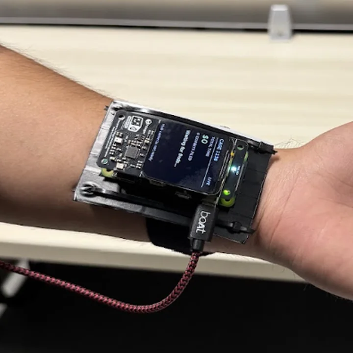 The wrist unit from the side: a forearm held level, palm up, a small lit screen on a black mount strapped across the inner wrist, a braided cable trailing from it.